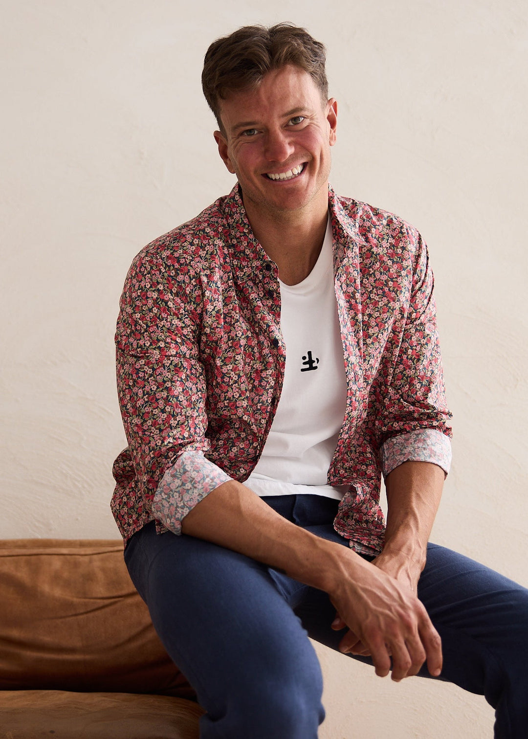 Man sitting on a brown couch wearing a patterned shirt and blue jeans against a plain wall.