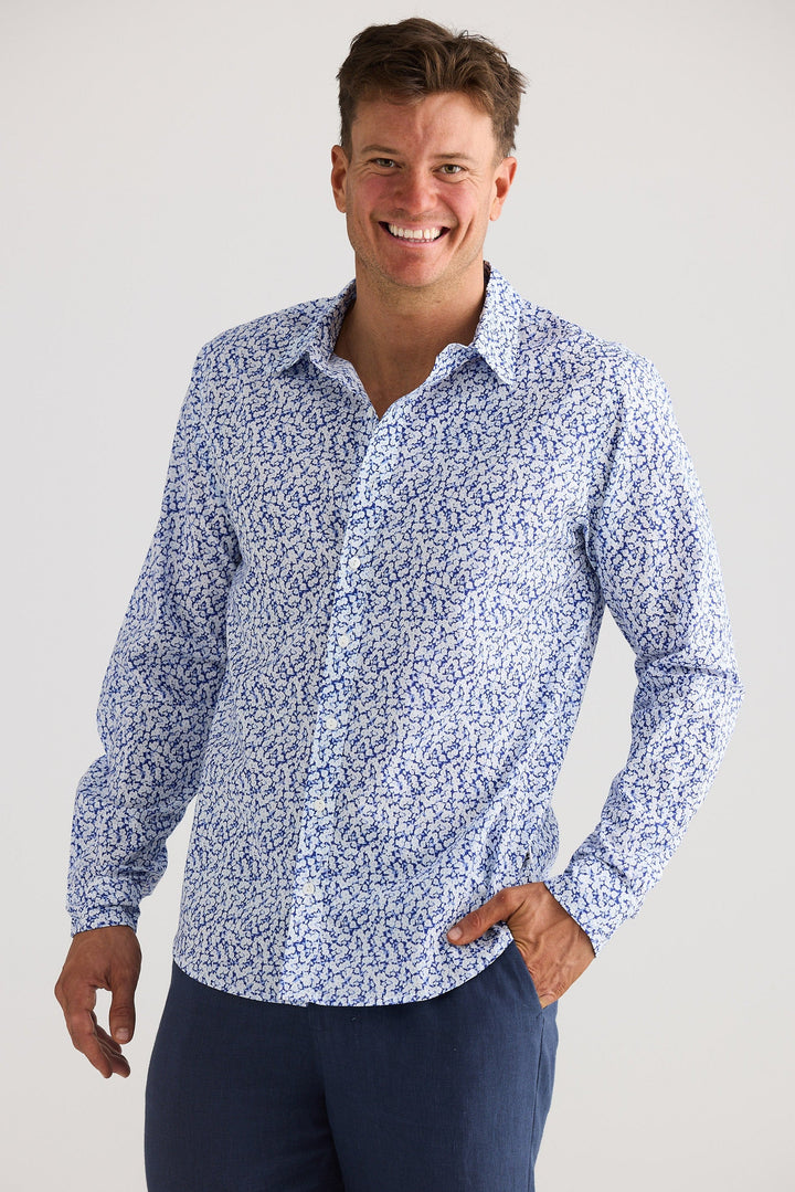 Man wearing a blue patterned shirt and navy pants against a white background