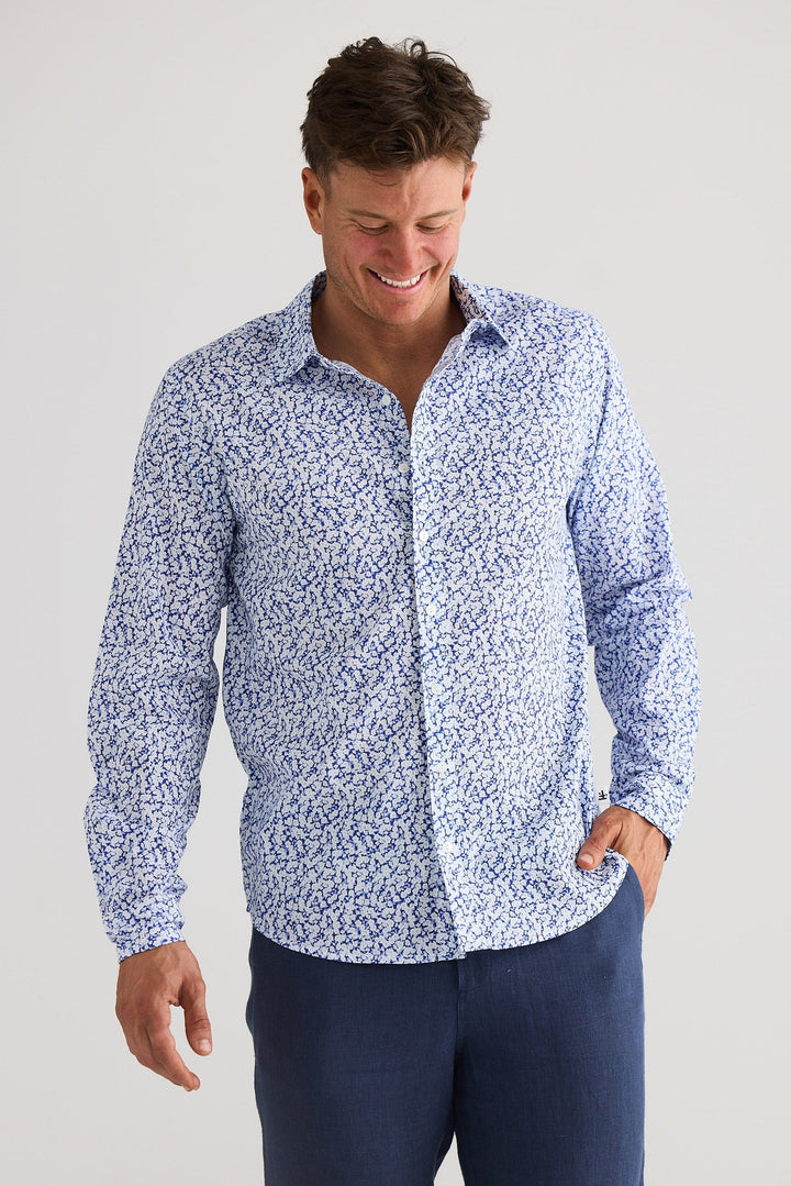 Man wearing a blue patterned shirt and dark pants on a light gray background