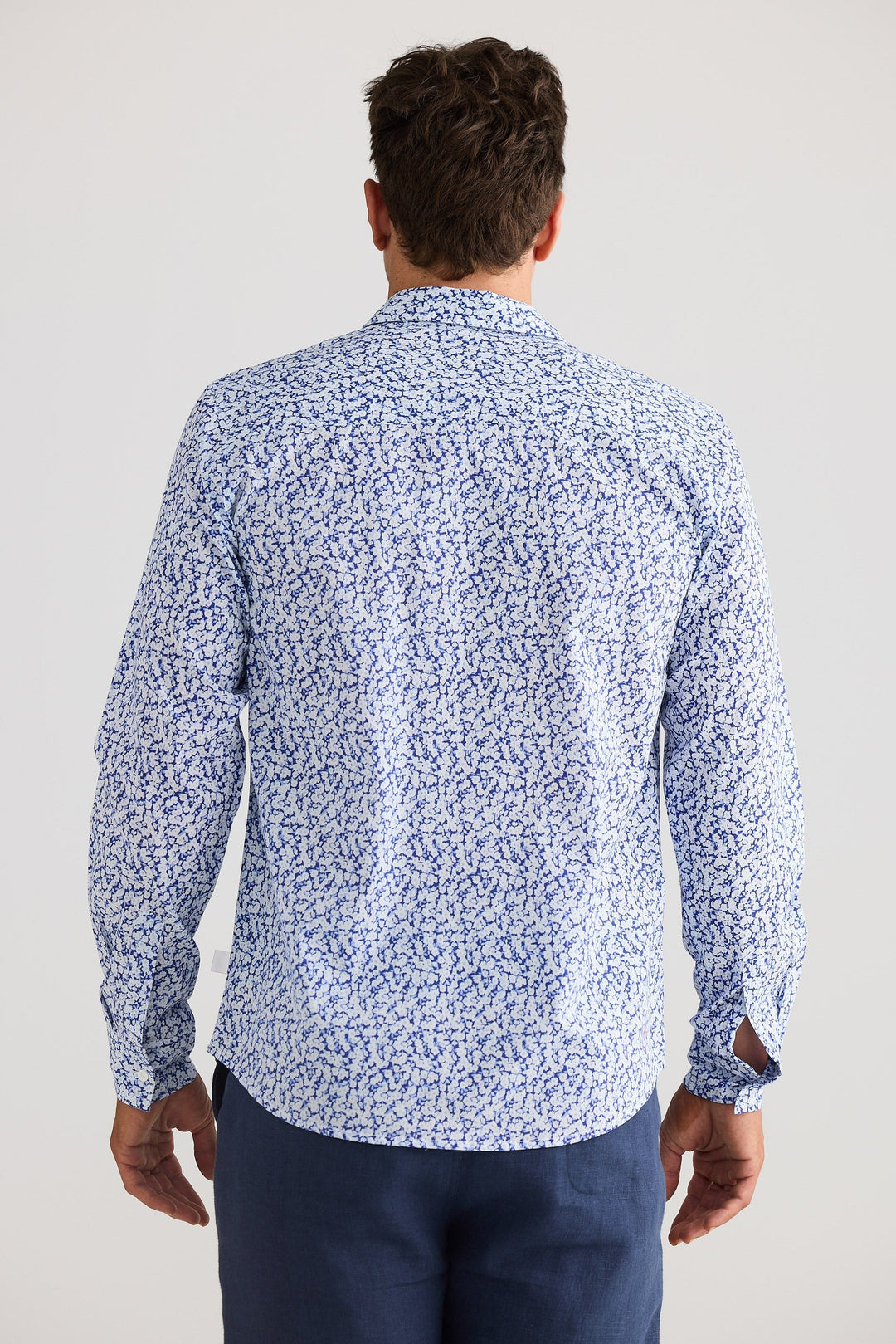 Man wearing a blue patterned shirt and dark pants on a white background