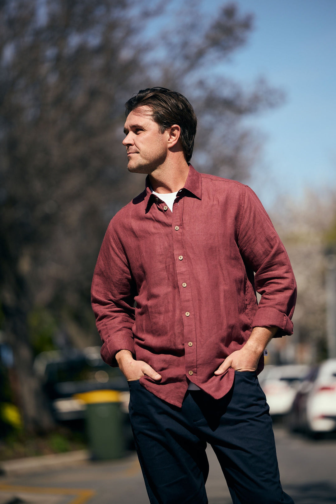 Man wearing a red shirt standing outdoors with trees and cars in the background