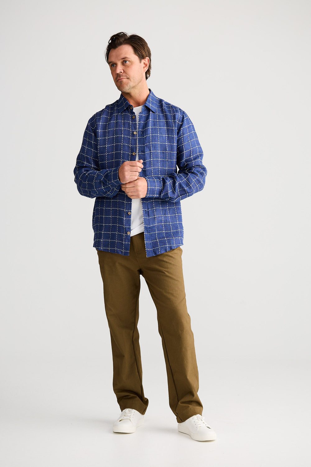 Man wearing a blue checkered shirt, white t-shirt, brown pants, and white sneakers on a white background