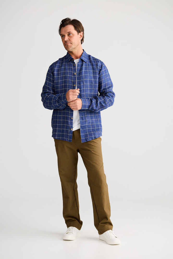 Man wearing a blue checkered shirt, white t-shirt, brown pants, and white sneakers on a white background