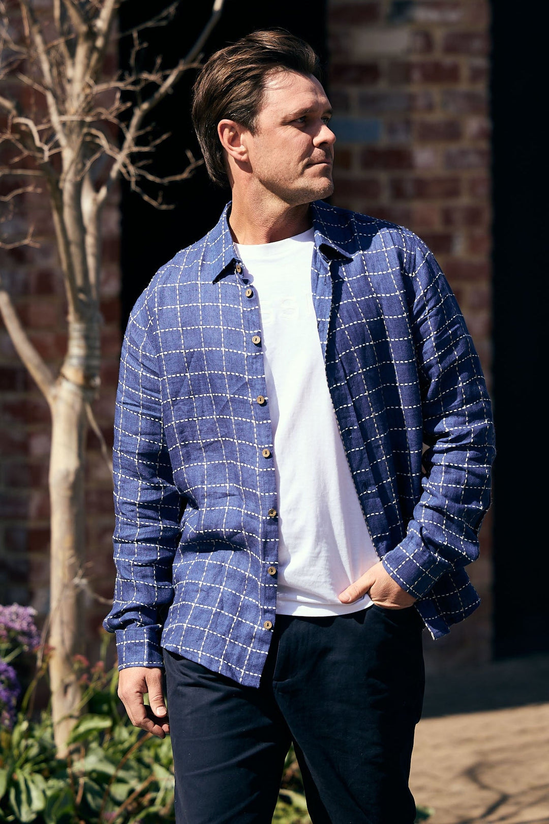 Man wearing a blue plaid shirt over a white t-shirt with black pants, standing outdoors.
