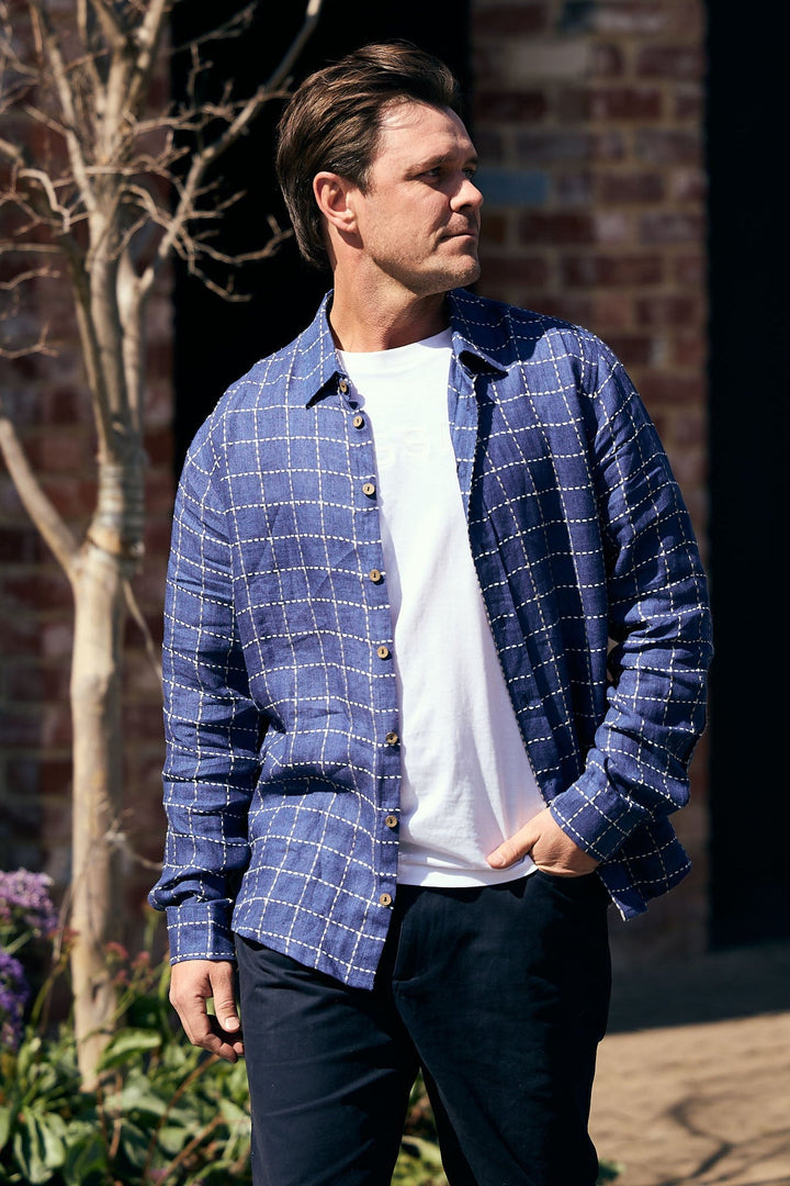 Man wearing a blue plaid shirt over a white t-shirt with black pants, standing outdoors.