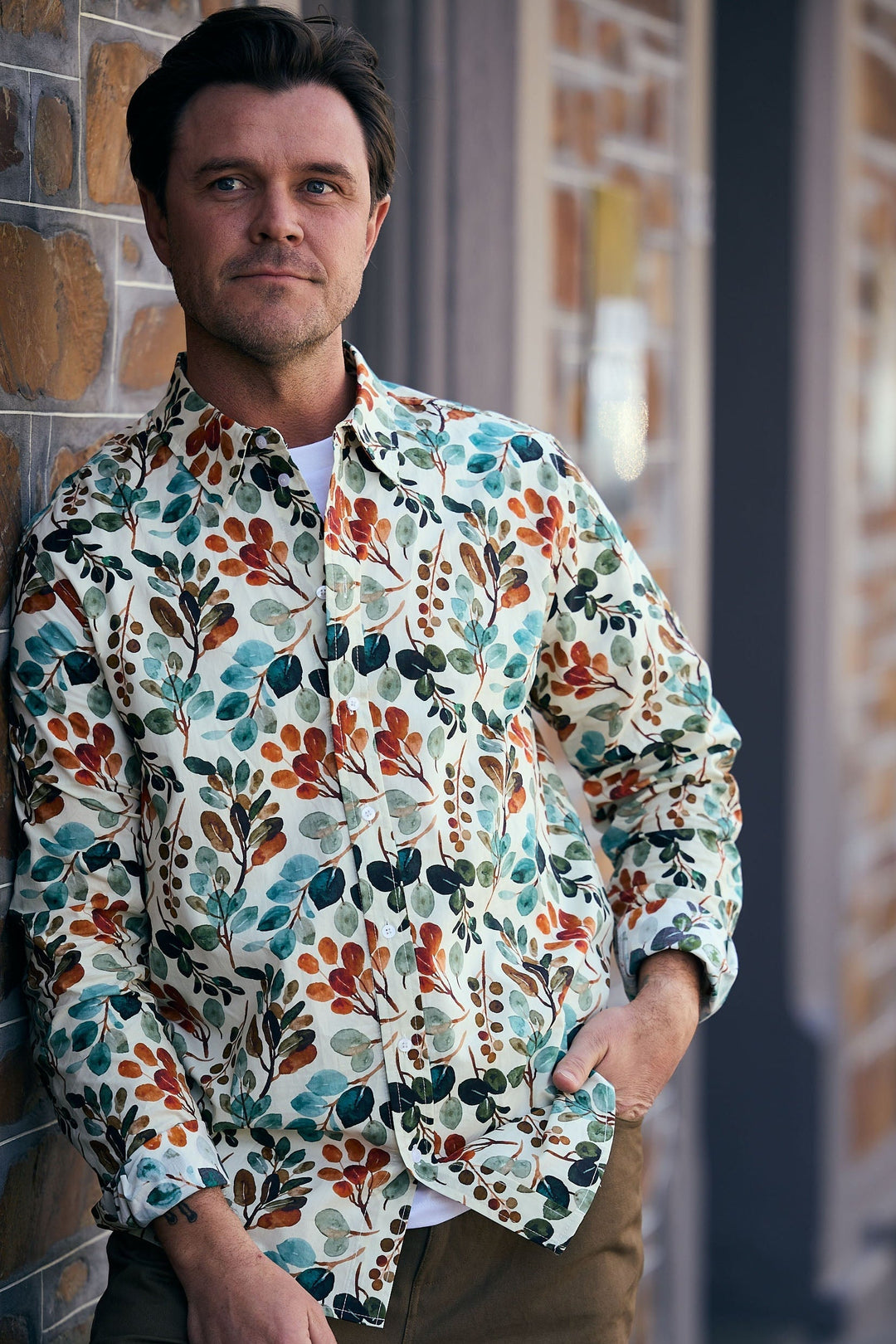 Man wearing a colorful floral shirt leaning against a brick wall.
