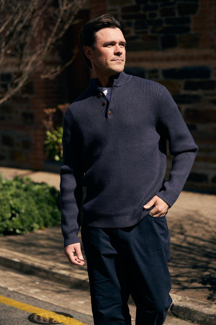 Man wearing a dark blue sweater and pants standing outdoors against a brick wall.