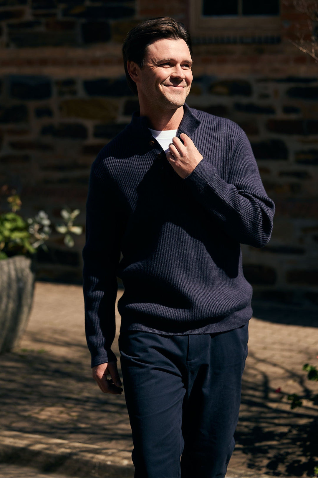 Man in a navy sweater and pants standing in front of a brick wall with plants.