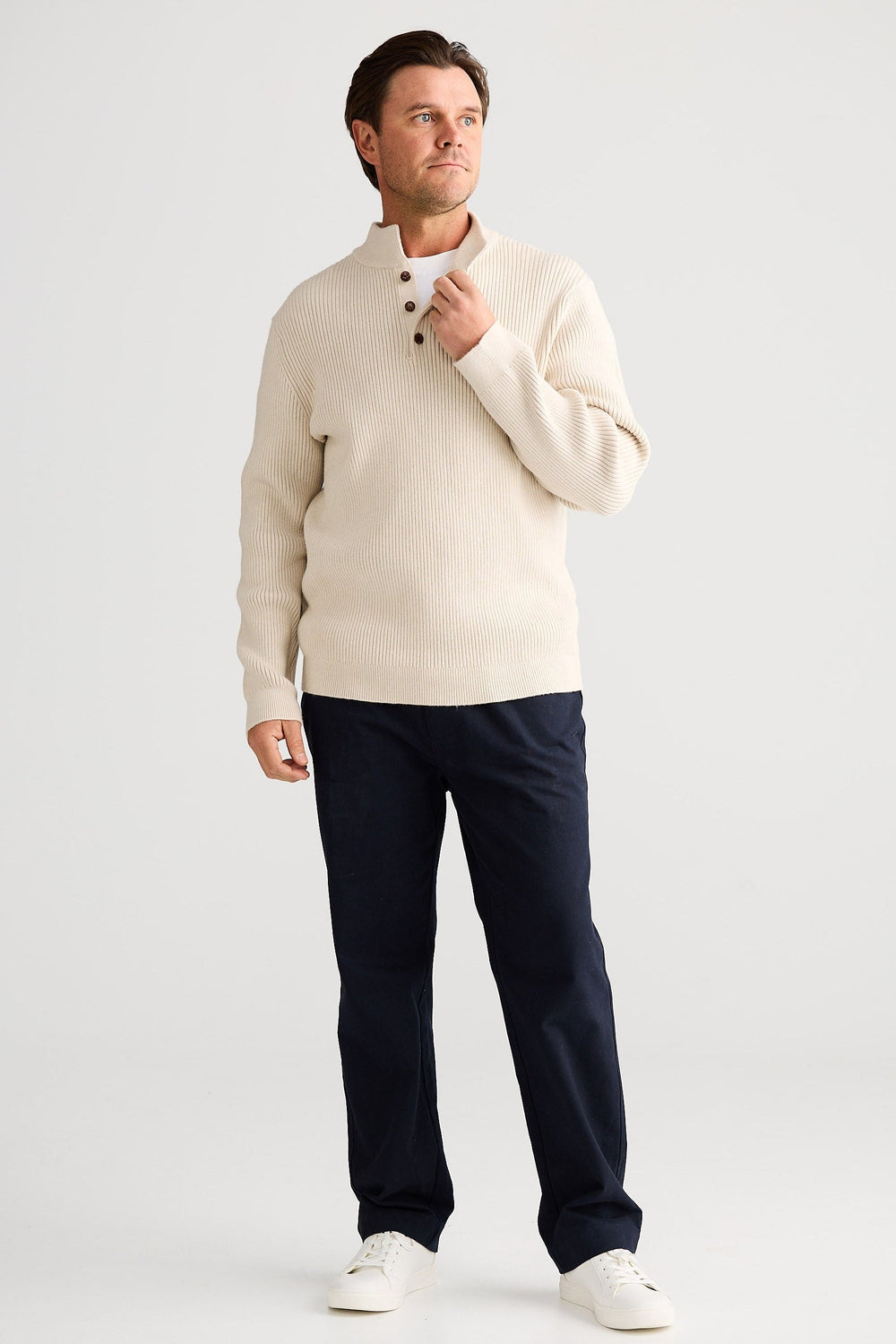 Man wearing a cream sweater and navy pants holding a coffee cup on a white background