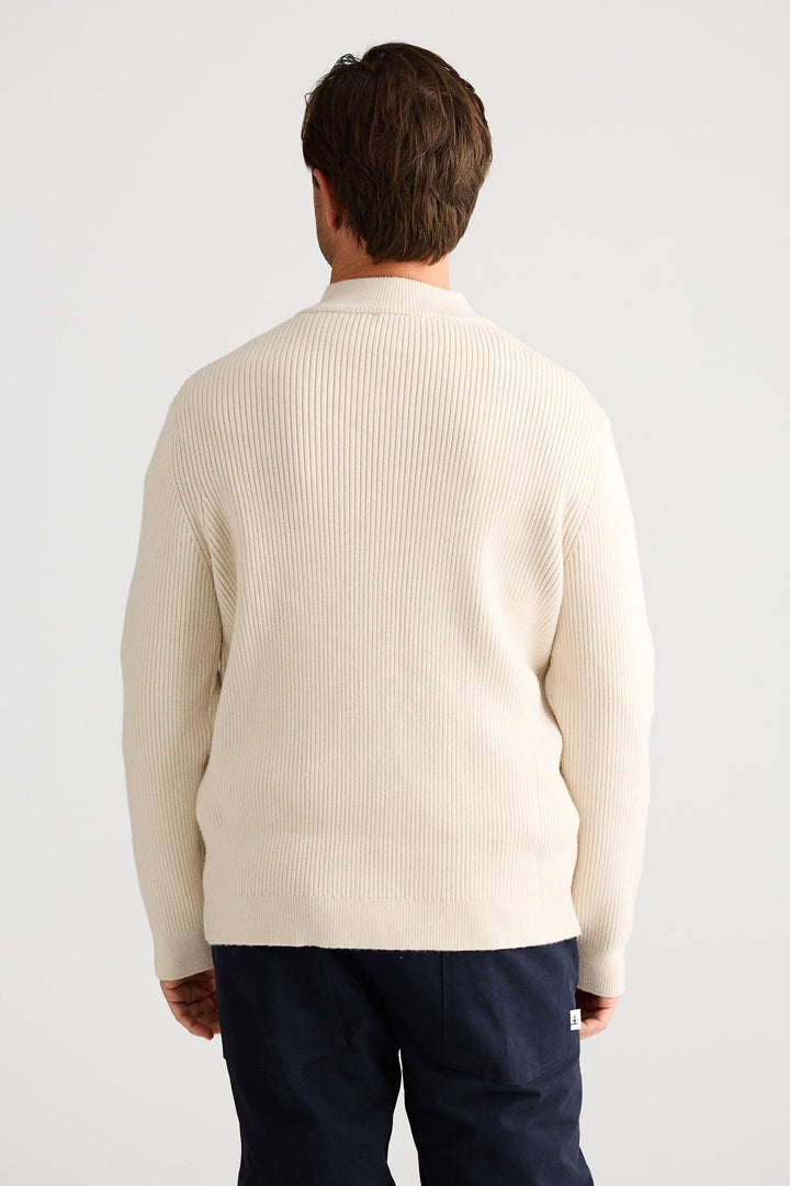 Man wearing a cream sweater and navy pants against a white background