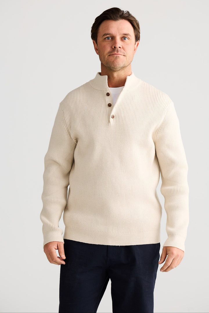 Man wearing a cream cardigan over a white shirt with navy pants on a light gray background