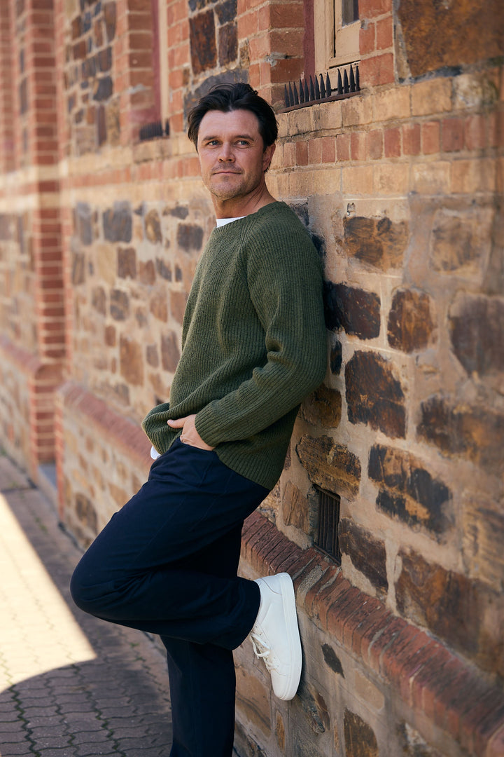 Man leaning against a brick wall wearing a green sweater and dark pants.