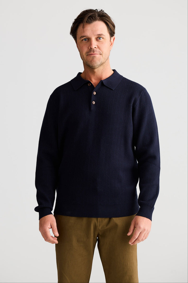 Man wearing a navy sweater over a collared shirt with brown pants on a light gray background