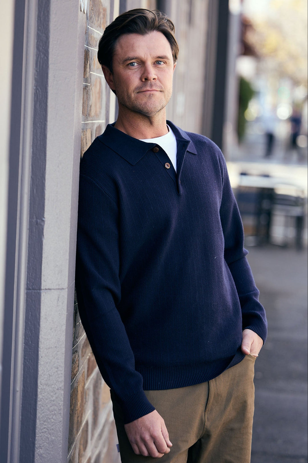 Man wearing a navy sweater leaning against a brick wall outdoors.