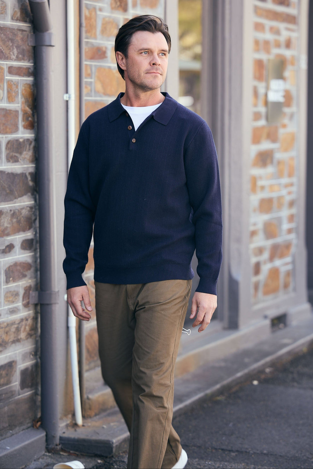 Man wearing a navy sweater and brown pants standing against a brick wall.