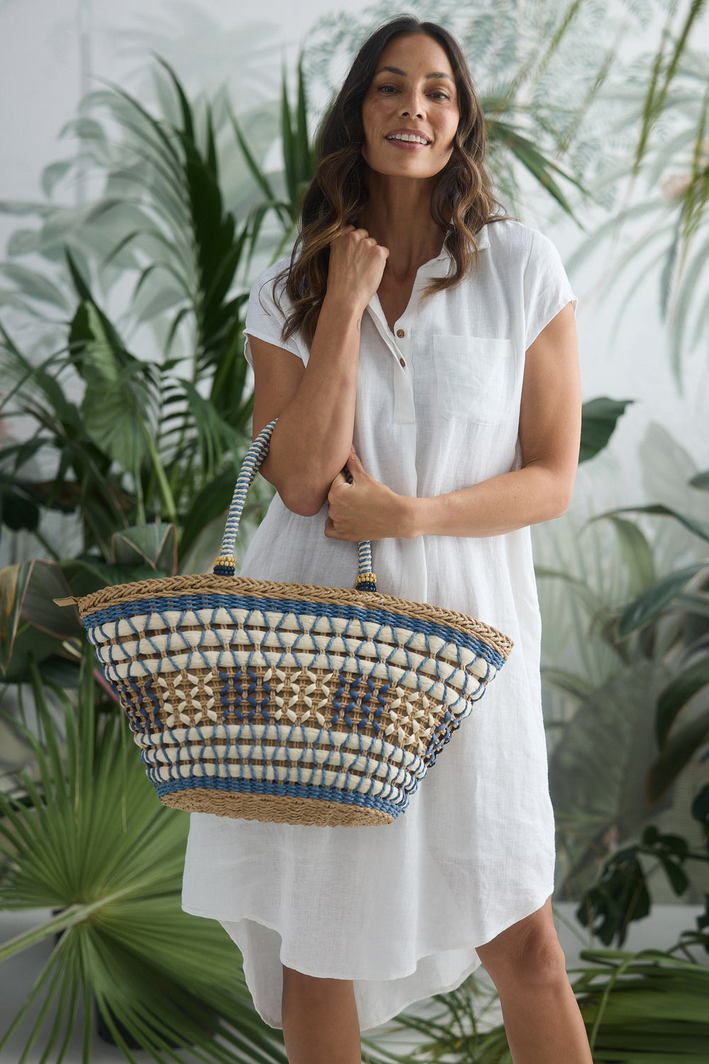 Woman in a white dress holding a woven bag with plants in the background
