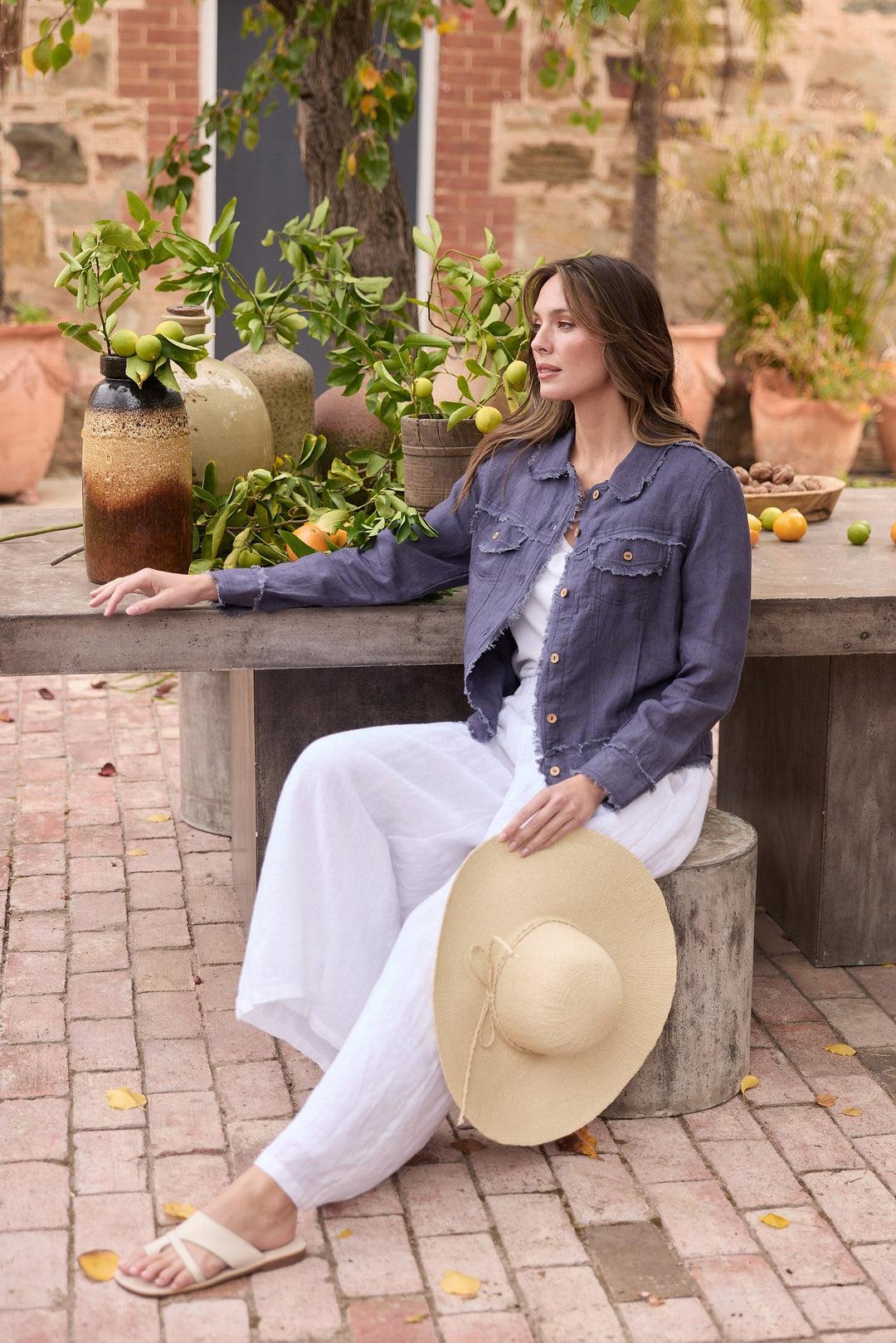 Woman sitting outdoors with a hat, surrounded by plants and fruit
