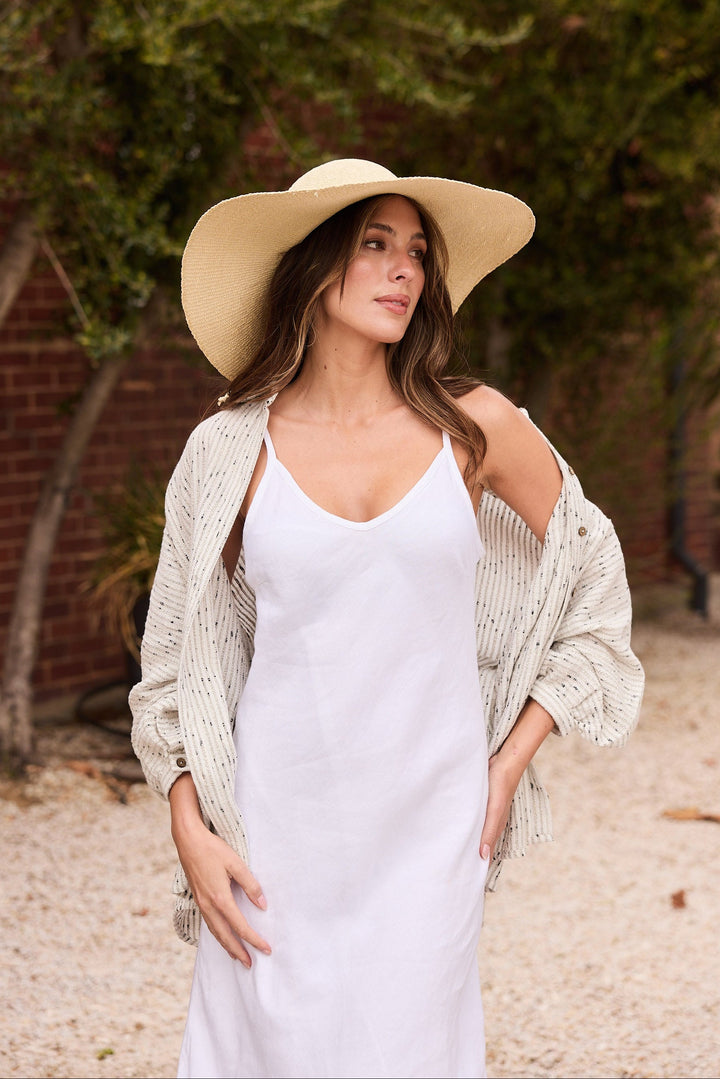 Woman wearing a white dress and beige hat outdoors with greenery in the background