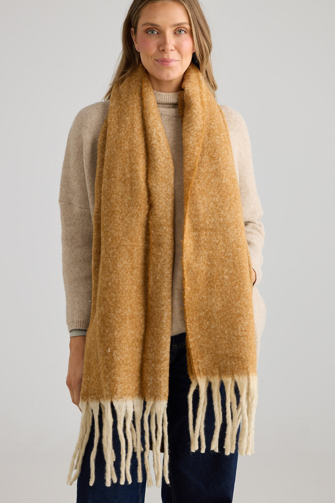 Woman wearing a mustard yellow scarf with white fringes on a plain background