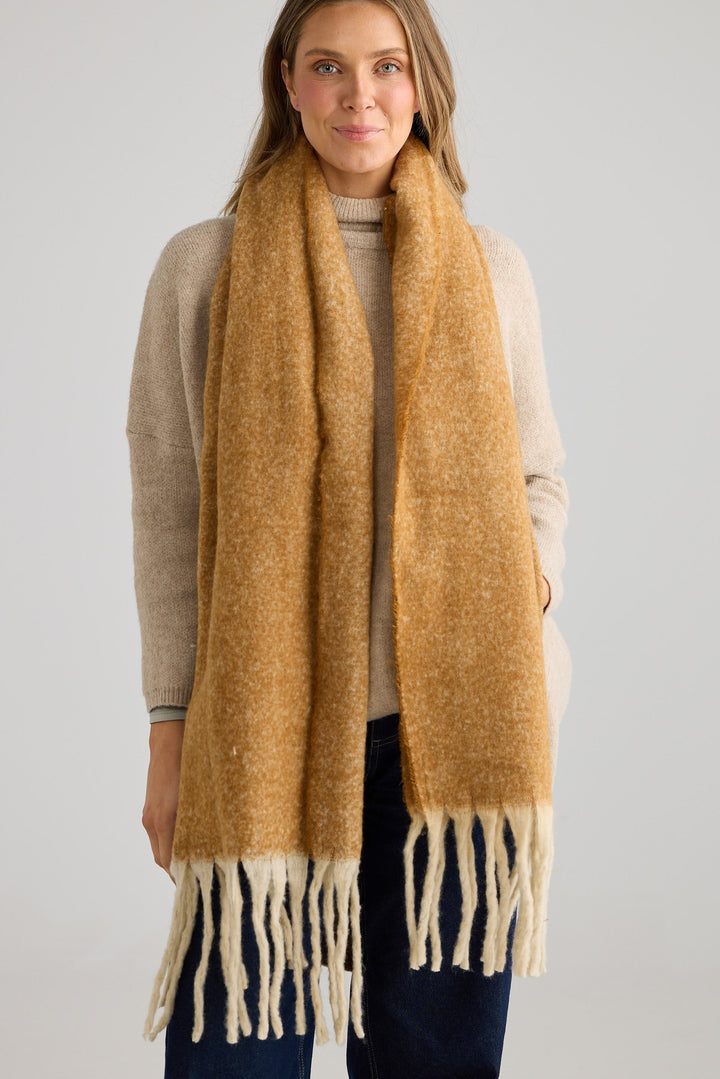 Woman wearing a mustard yellow scarf with white fringes on a plain background