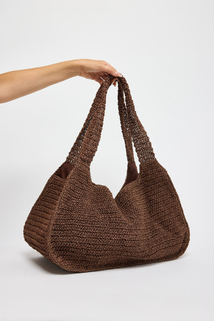 Large slouchy hobo bag in chunky chocolate brown woven raffia with braided rope handles, held by hand against a white background
