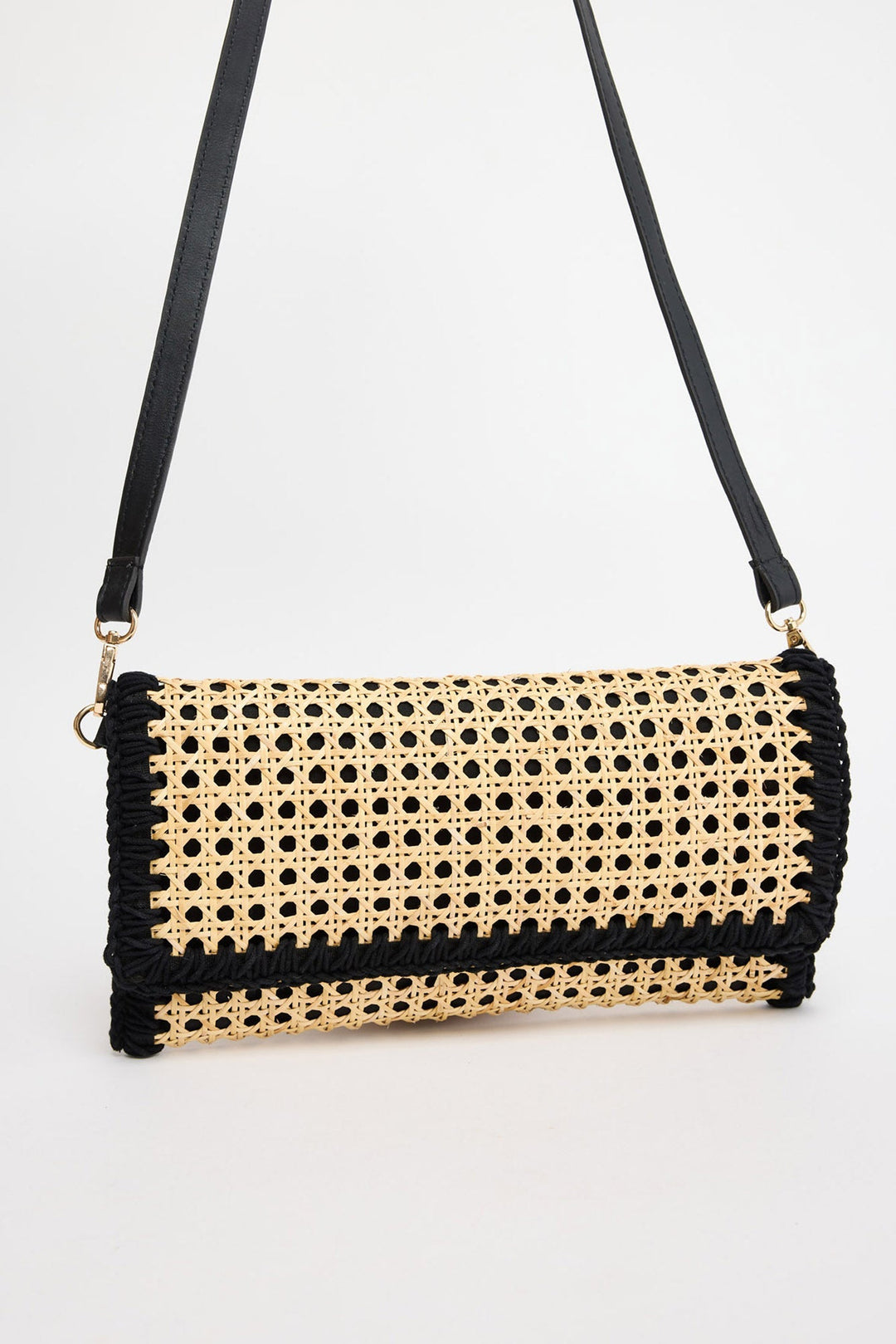 Rectangular fold-over crossbody bag in natural rattan cane weave with black whipstitched trim along all edges and a black leather adjustable strap with gold-tone hardware, shown against a white background