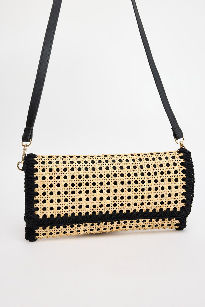 Rectangular fold-over crossbody bag in natural rattan cane weave with black whipstitched trim along all edges and a black leather adjustable strap with gold-tone hardware, shown against a white background