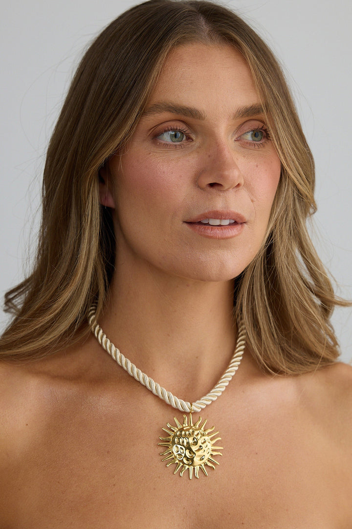 Woman wearing a gold necklace with a sun pendant against a plain background