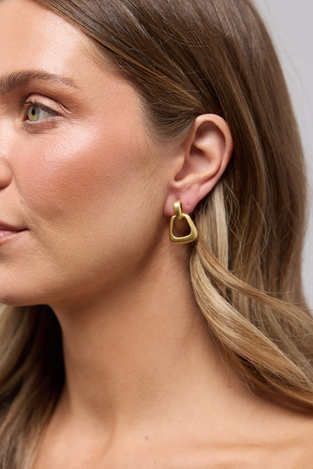 Swell Earrings in Gold worn on the ear, showing the rounded-triangle geometric drop against the lobe.