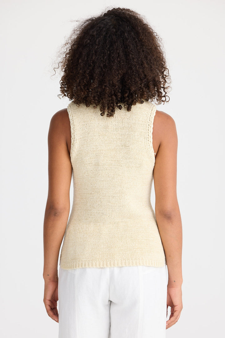 Person wearing a beige sleeveless top and white pants on a plain background