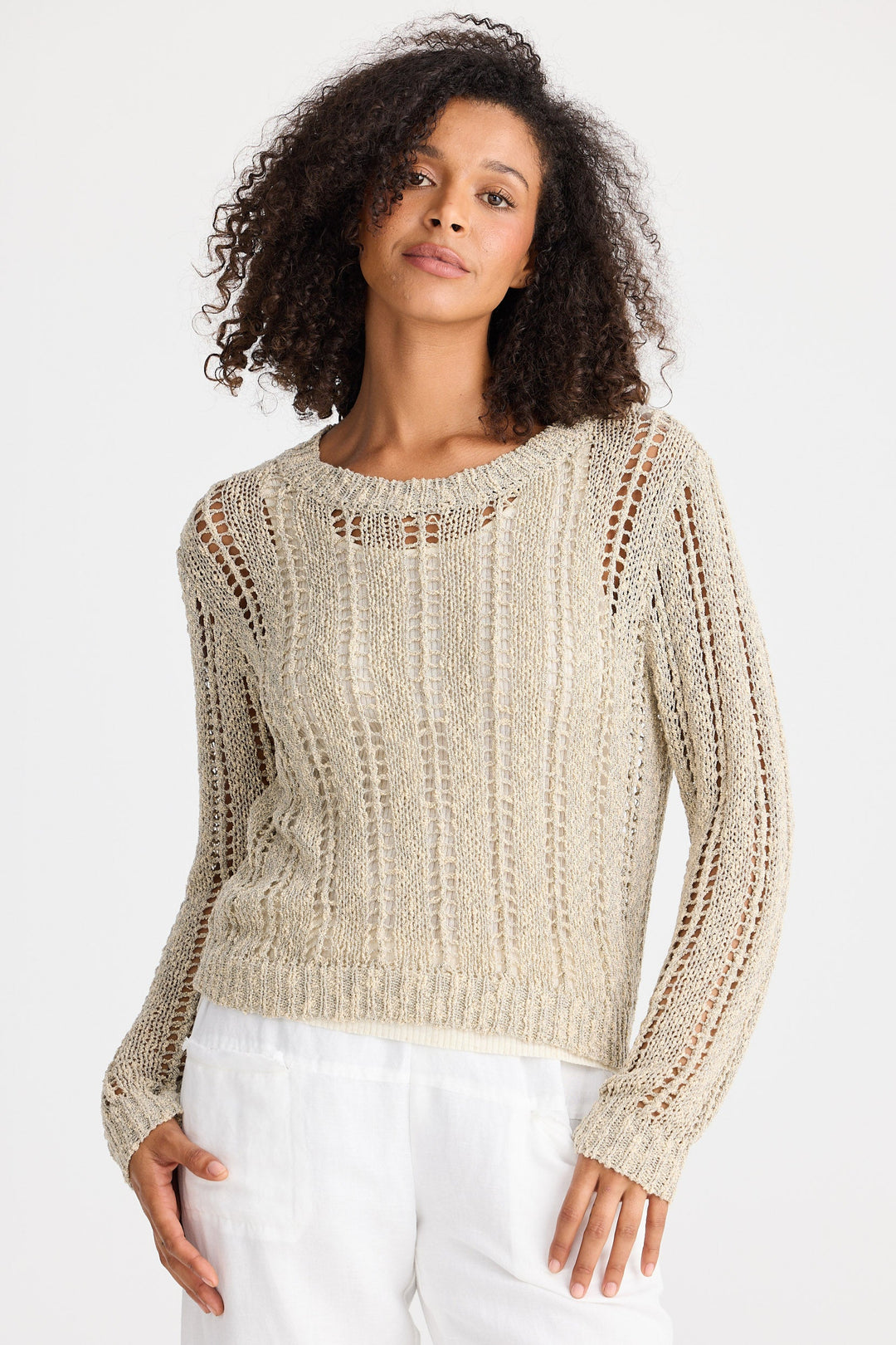 Woman wearing a beige knit sweater and white pants on a plain background