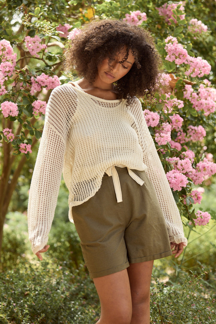 Woman wearing a cream knit sweater and green shorts standing in a garden with pink flowers.