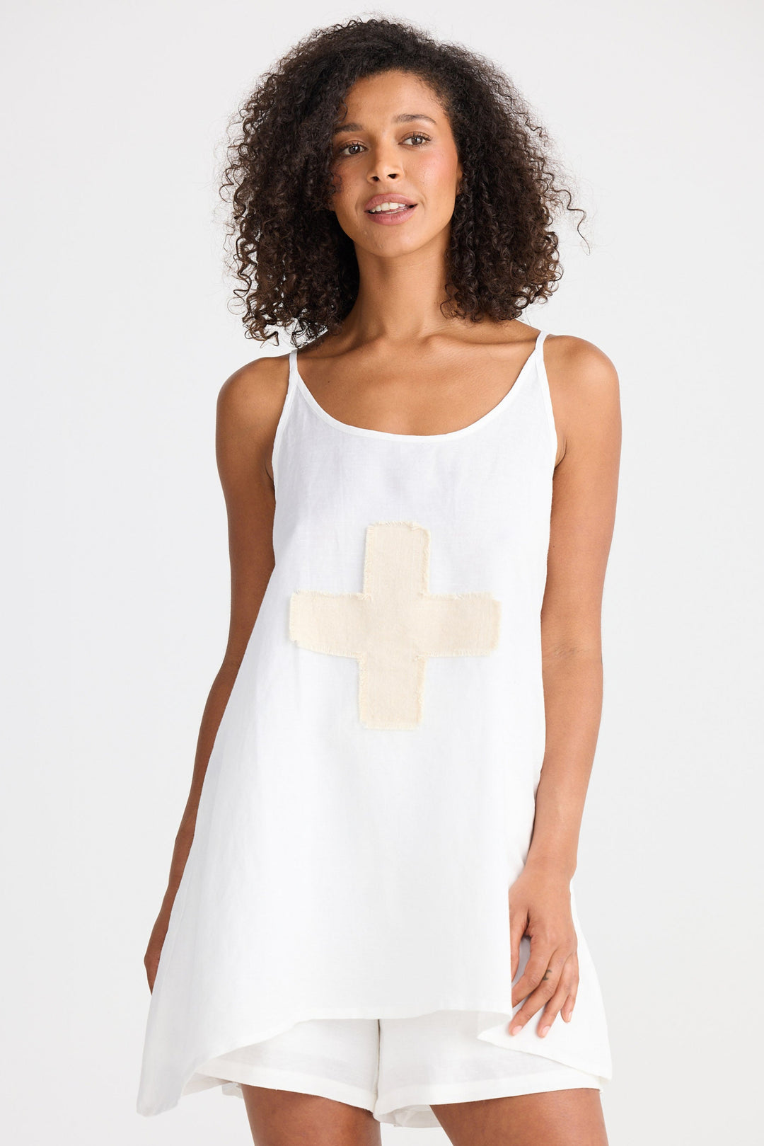Woman wearing a white sleeveless top with a cream cross design on a plain background