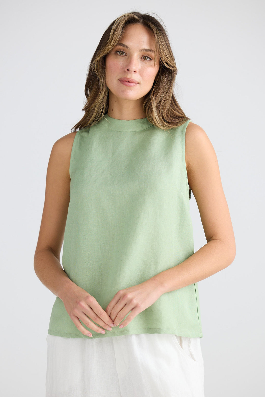 Woman wearing a light green sleeveless top and white pants on a plain background