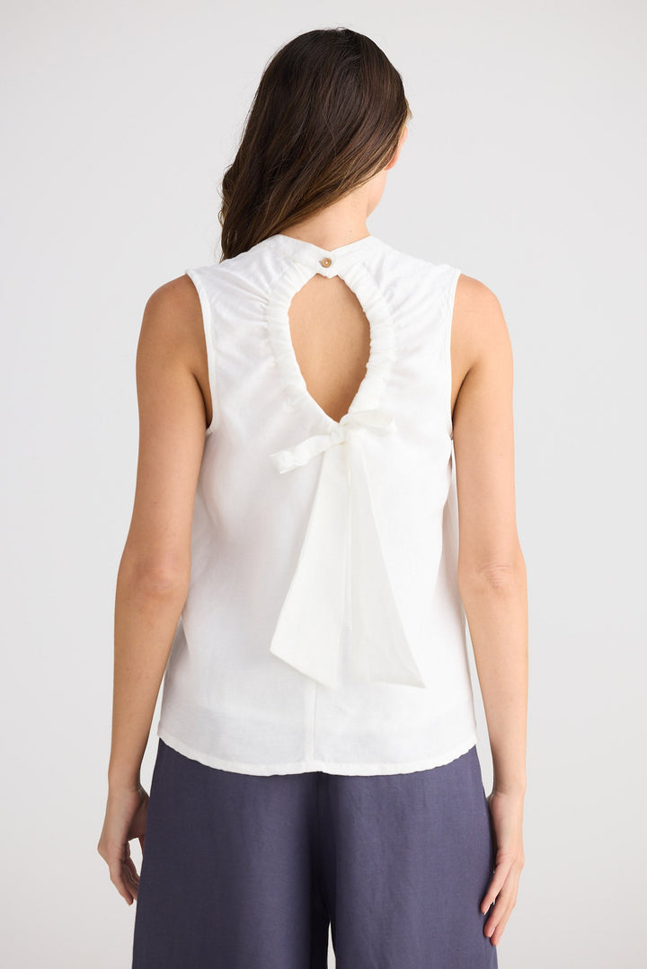 Woman wearing a white sleeveless top with a bow detail on the back, standing against a plain background.