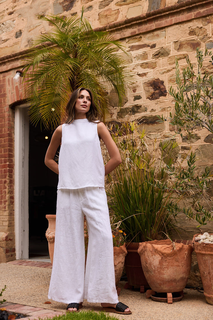 Woman in a white outfit standing in front of a stone wall with plants.