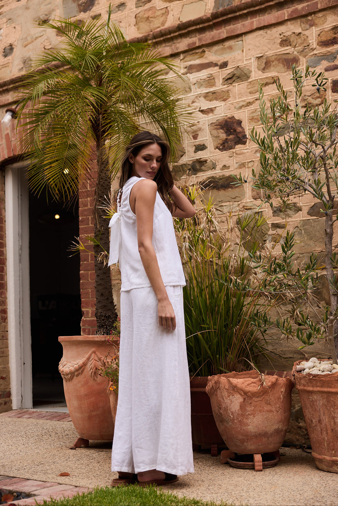 Woman in a white dress standing in front of a stone wall with potted plants.
