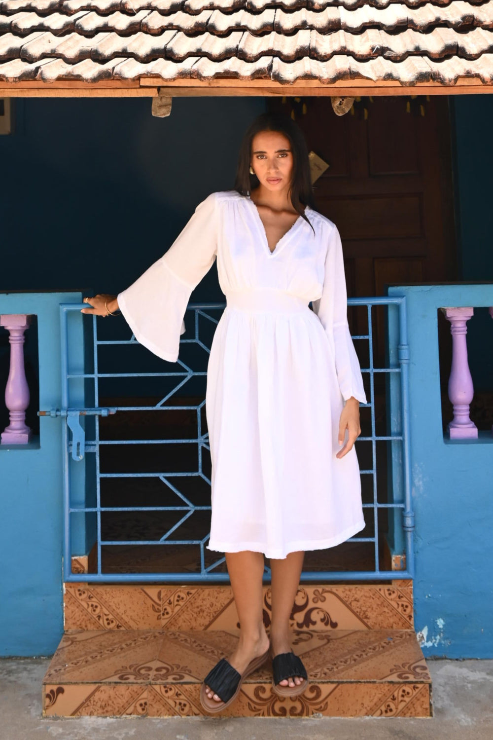 Woman in a white dress standing in front of a blue wall and palm trees in the background.