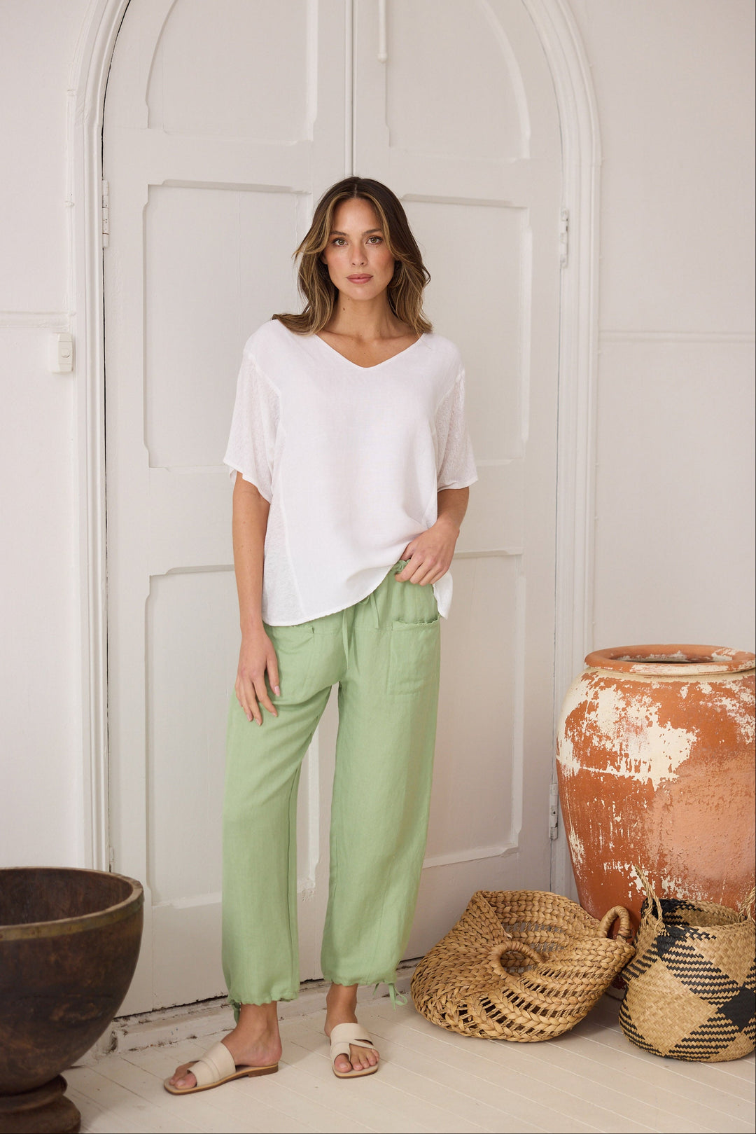 Woman wearing a white top and green pants standing in a room with decorative pots and baskets.