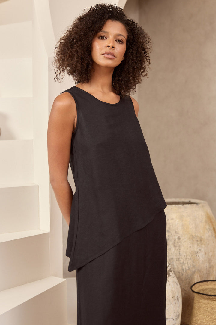 Woman wearing a black sleeveless top and skirt standing in a minimalistic room with white shelves and a neutral wall.
