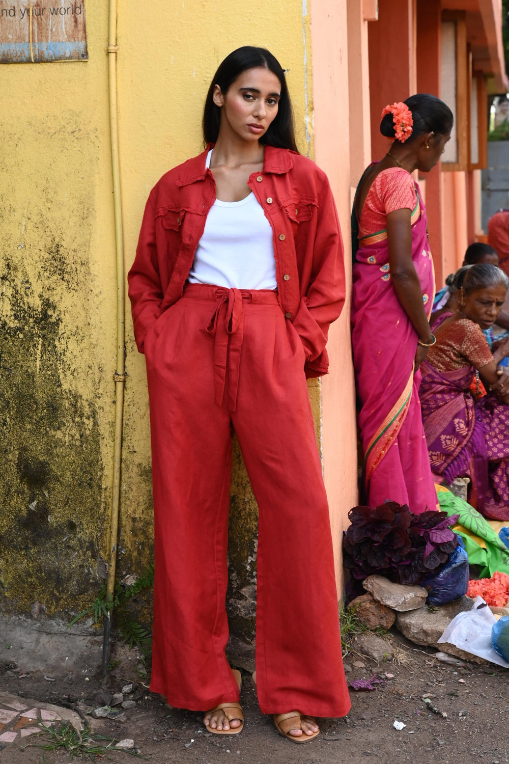 A woman standing against a yellow textured wall wearing a red jacket and pants and a white top.