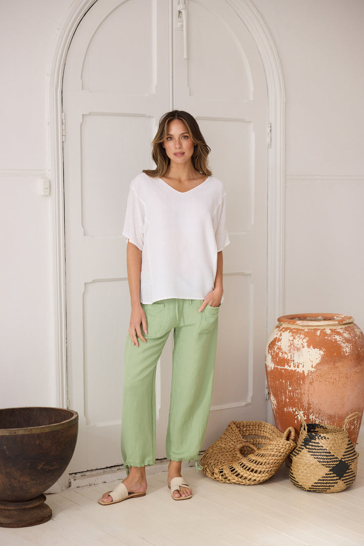 Woman wearing a white top and light green pants standing in a room with decorative pots and a door.