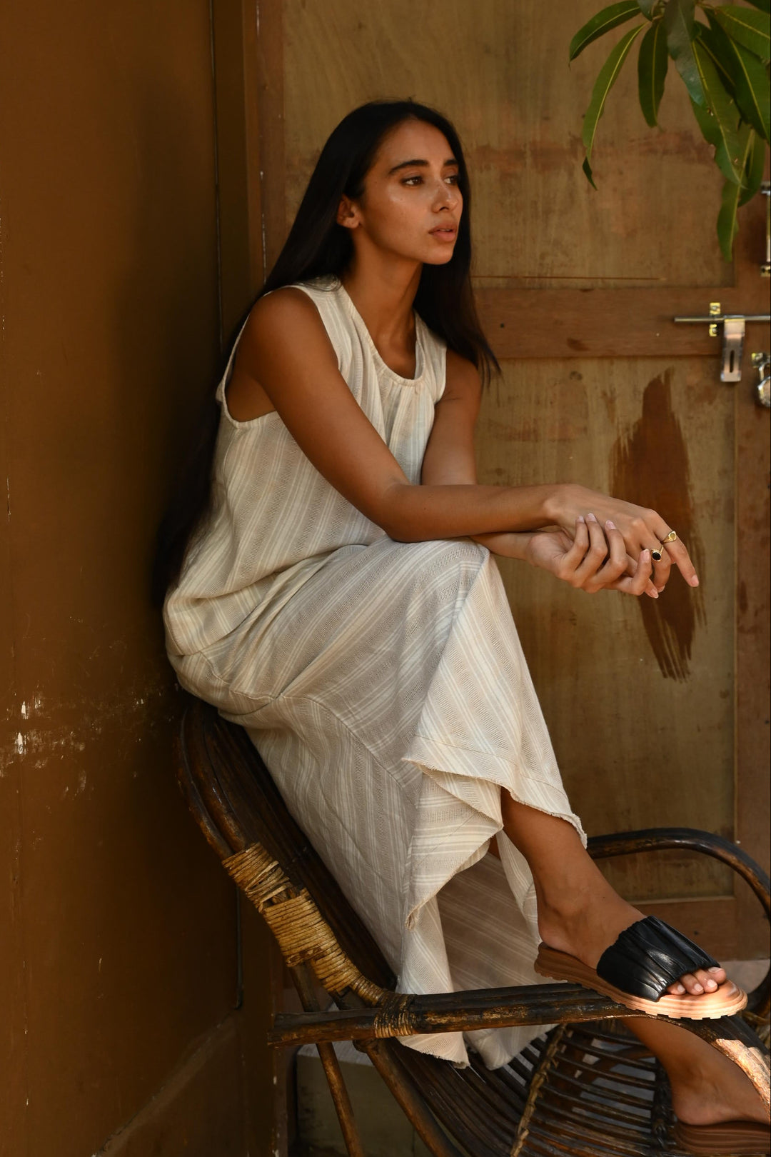 Woman in a light coloured striped top and skirt set sitting on a wooden chair with a plant in the corner.