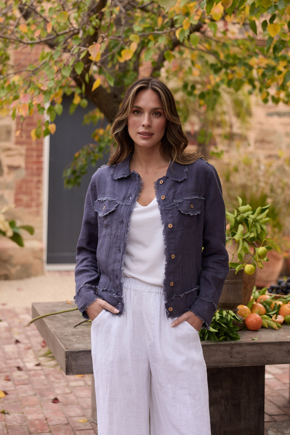 Woman standing outdoors near a wooden table with fruits, wearing a navy jacket and white pants.