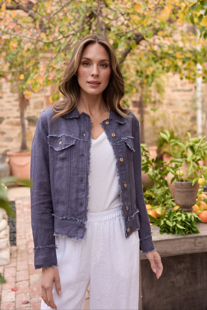 Woman wearing a navy jacket and white pants standing outdoors with trees and plants in the background