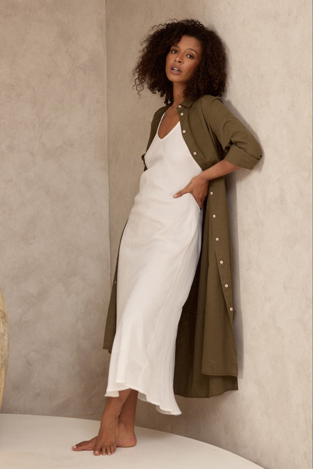 Woman in a white dress and green shirt dress standing against a textured wall with large ceramic pots.