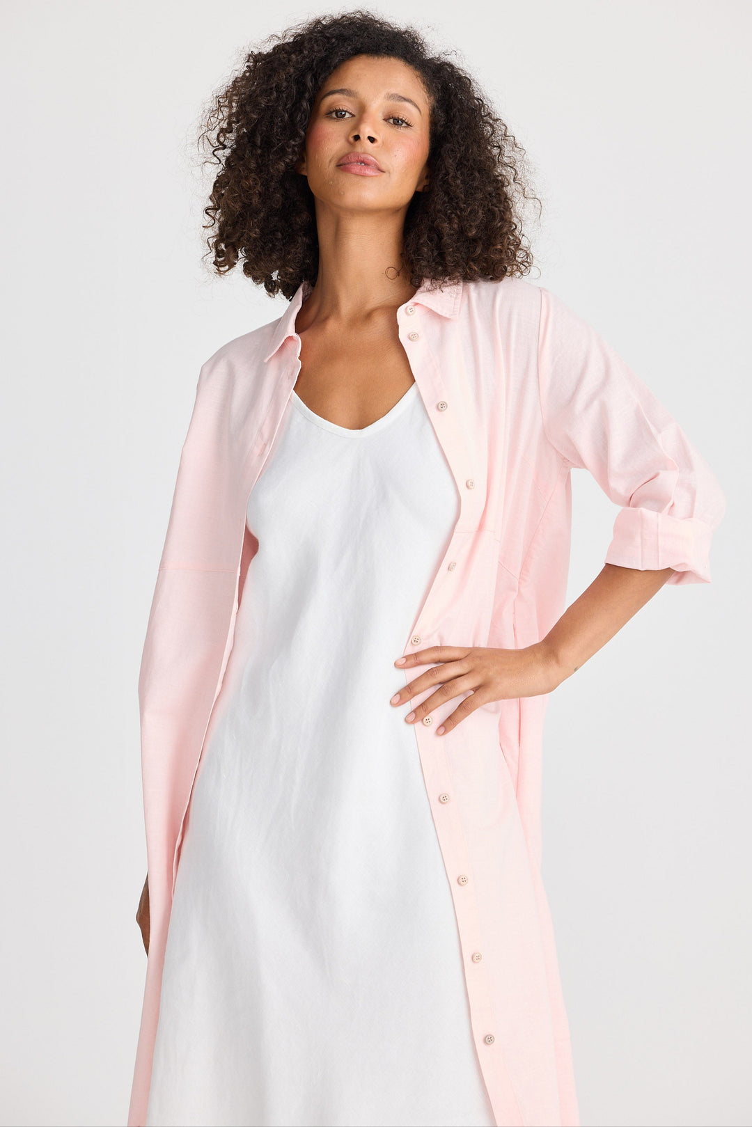 Woman wearing a light pink shirt dress open over a white dress on a plain background