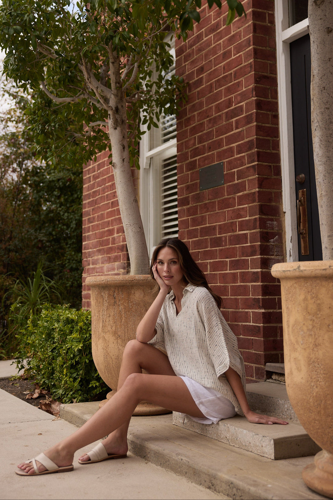 Woman sitting on steps outside a brick building with trees and plants around.