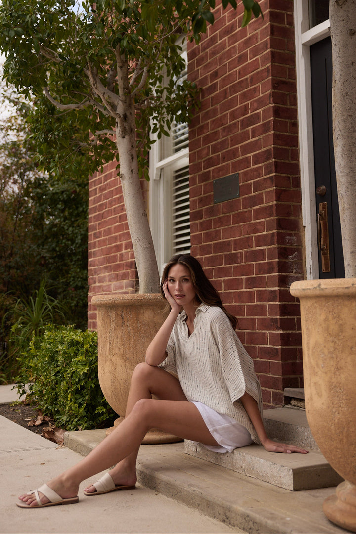 Woman sitting on steps outside a brick building with trees and plants around.