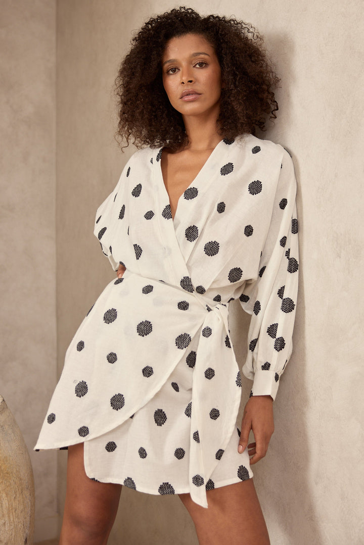 Woman wearing a white dress with black polka dots against a beige wall.